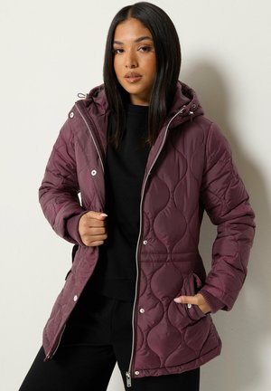 Femme portant une veste matelassée à capuche couleur prune sur une tenue noire, une main dans la poche et l'autre tenant la fermeture éclair de la veste.