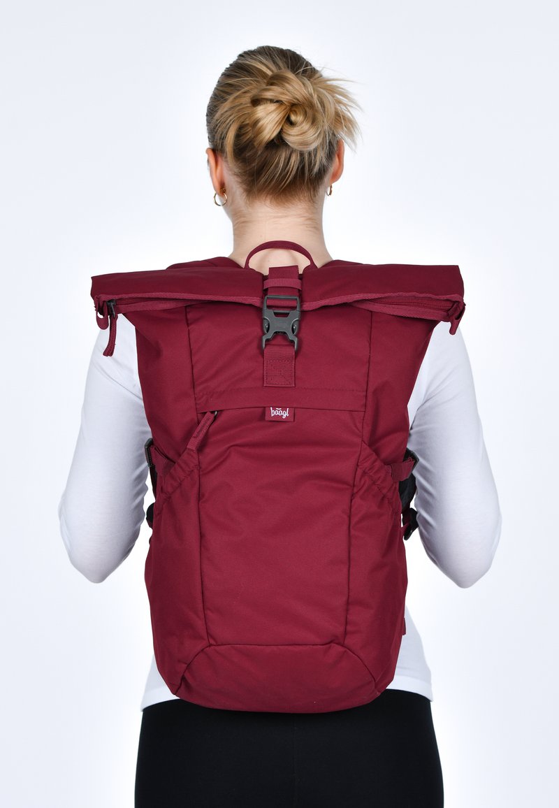 Zaino in tessuto rosso con chiusura a rotolo e spallacci regolabili. Presenta una fibbia nera e tasche laterali, progettato per utilità e comfort.