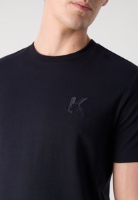 T-shirt en coton noir avec un col rond et un logo "K" brodé ton sur ton sur la poitrine gauche. Texture lisse, coupe ajustée.