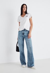 Mladá žena s dlhými tmavými vlasmi, oblečená v bielej tričku Levi's, vo voľných svetlomodrých džínsach, s bielymi teniskami a malou čiernou kabelkou.