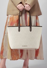 Biała torba tote z wytłaczanym wzorem, brązowymi skórzanymi uchwytami i metalicznym akcentem logo. Stylizowana z beżową kurtką i wielokolorową spódnicą.