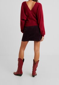 Femme portant un pull rouge profond à dos en V, une mini jupe sombre, des bottes de cowboy rouges mi-mollet et un collier argenté avec un pendentif dans le dos.