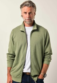 Maglioncino verde chiaro con zip, texture a coste, maniche lunghe e colletto alto. Indossato sopra una camicia bianca, abbinato a jeans scuri.
