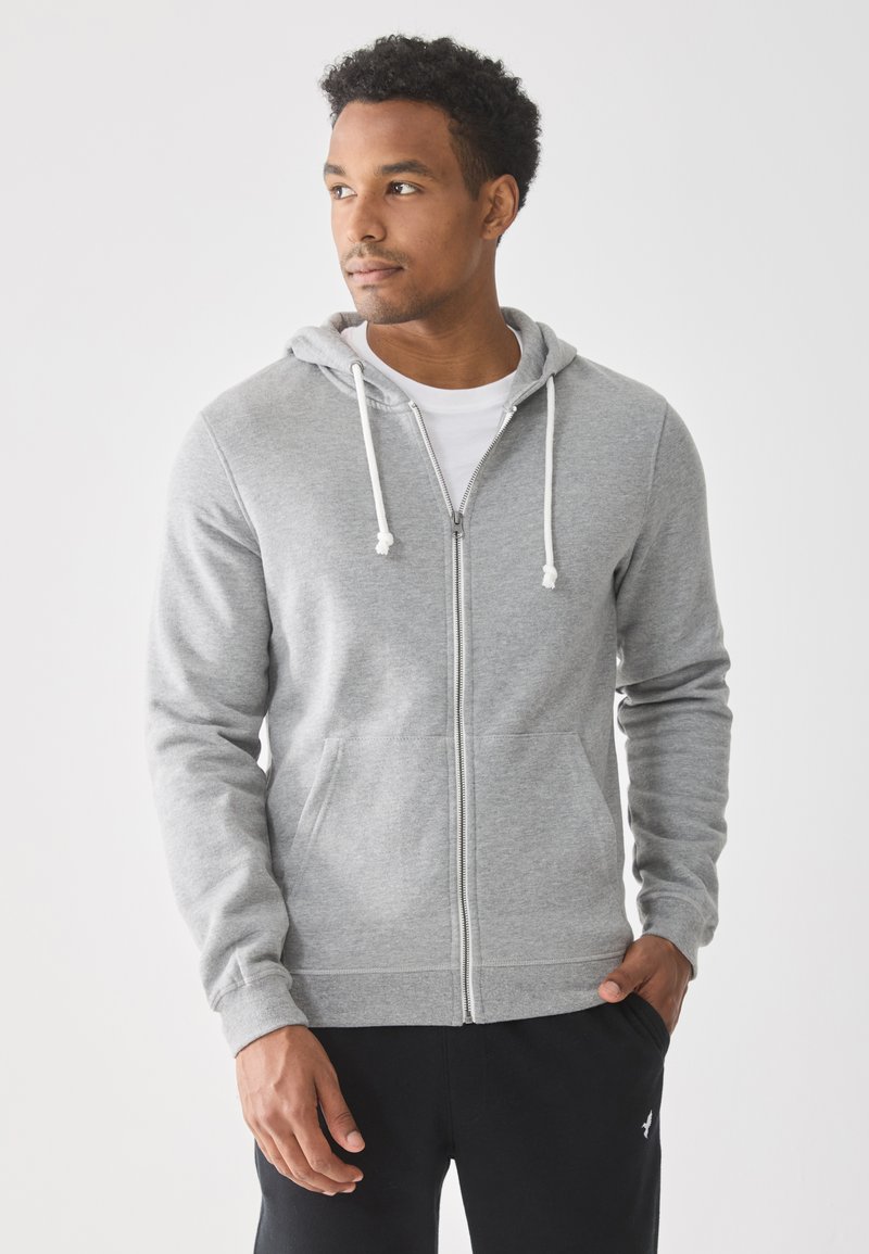 Mann in heller grauer Zip-Up-Hoodie und schwarzen Hosen, steht mit einer Hand in der Tasche und schaut nach links vor einem weißen Hintergrund.