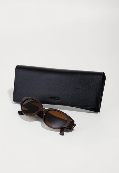 Bruine zonnebril met donkere lenzen, hoekig frame en gegraveerde accenten, neergelegd naast een zwart leren etui met een gladde textuur.