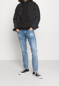 Černá denimová bunda přes černou mikinu, doplněná o světle modré džíny a černé tenisky. Tento outfit se vyznačuje jednoduchými texturami a ležérním designem.