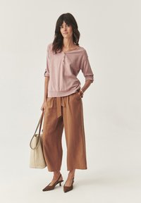 Maglietta a maniche lunghe rosa chiaro con bottoni, abbinata a pantaloni larghi marroni e décolleté marroni con cinturino. Accessoriata con una borsa tote beige.