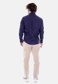 Camicia a maniche lunghe blu navy con bottoni, abbinata a pantaloni beige chiaro e sneakers bianche, mostrata di spalle su uno sfondo bianco.