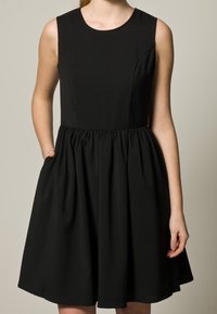 Robe noire sans manches avec un col rond, un corsage ajusté et une jupe évasée. Elle comporte des poches latérales et un tissu texturé.