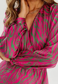 Mujer con un vestido rosa de mangas largas y rayas de cebra abstractas verdes, con la mano cerca de la cara, llevando un collar de cadena dorada.