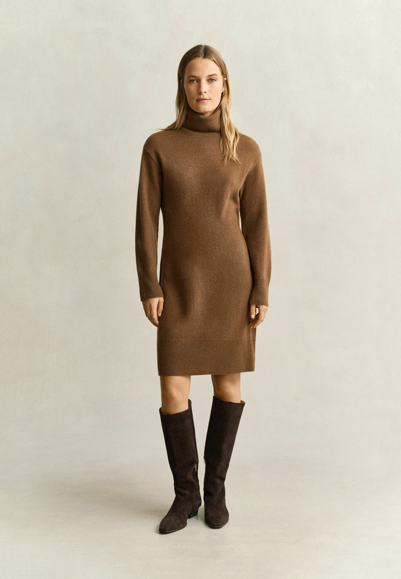 Femme debout portant une robe-pull marron à manches longues et col roulé avec des bottes marron foncé montant jusqu'au genou sur un fond clair uni.