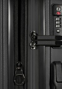 Primer plano de la cremallera de una maleta negra con tiradores de marca y un candado con combinación TSA sobre una superficie texturizada.