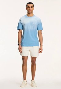 Jasnoniebieski t-shirt z krótkimi rękawami w gradiencie, z małym nadrukiem na piersi. Model nosi go w połączeniu z białymi szortami i trampkami.