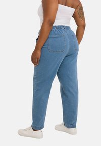 Pantalones de mezclilla ligero con cinturilla elástica, dos bolsillos traseros y piernas ajustadas, combinados con zapatillas blancas.