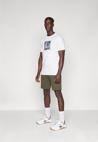 Camiseta blanca de manga corta con un diseño gráfico azul, combinada con pantalones cortos verdes y zapatillas blancas con detalles negros y una suela texturizada.