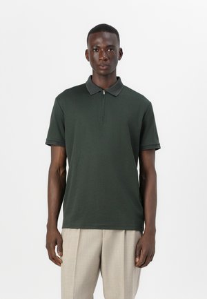 Dark green polo shirt with a zippered collar, short sleeves, and contrasting gray accents. Styled with light beige trousers.