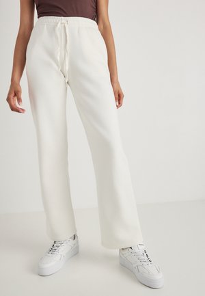 Hvite, vide joggebukser med elastisk linning og snøring. Glatt tekstur, sammen med hvite sneakers med teksturert såle.