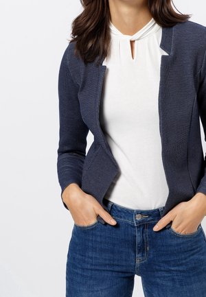 Woman wearing a navy textured blazer over a white keyhole-neck blouse and blue jeans with hands in pockets, against a plain background.