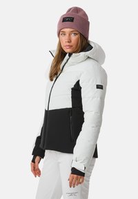 Giacca zip-up puffy bianca e nera con cappuccio, dotata di tasche laterali e polsini elasticizzati. Abbinata a un berretto a coste rosa.