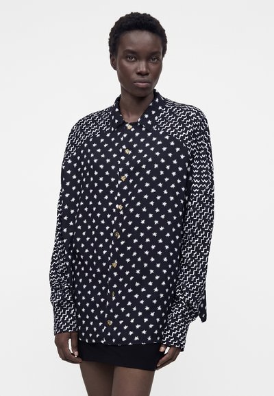 Νεαρή γυναίκα φοράει μαύρο πουκάμισο με κουμπιά με λευκά αστέρια και ζικ-ζακ σχέδια, σε συνδυασμό με μαύρη φούστα, στέκεται μπροστά σε απλό φόντο.