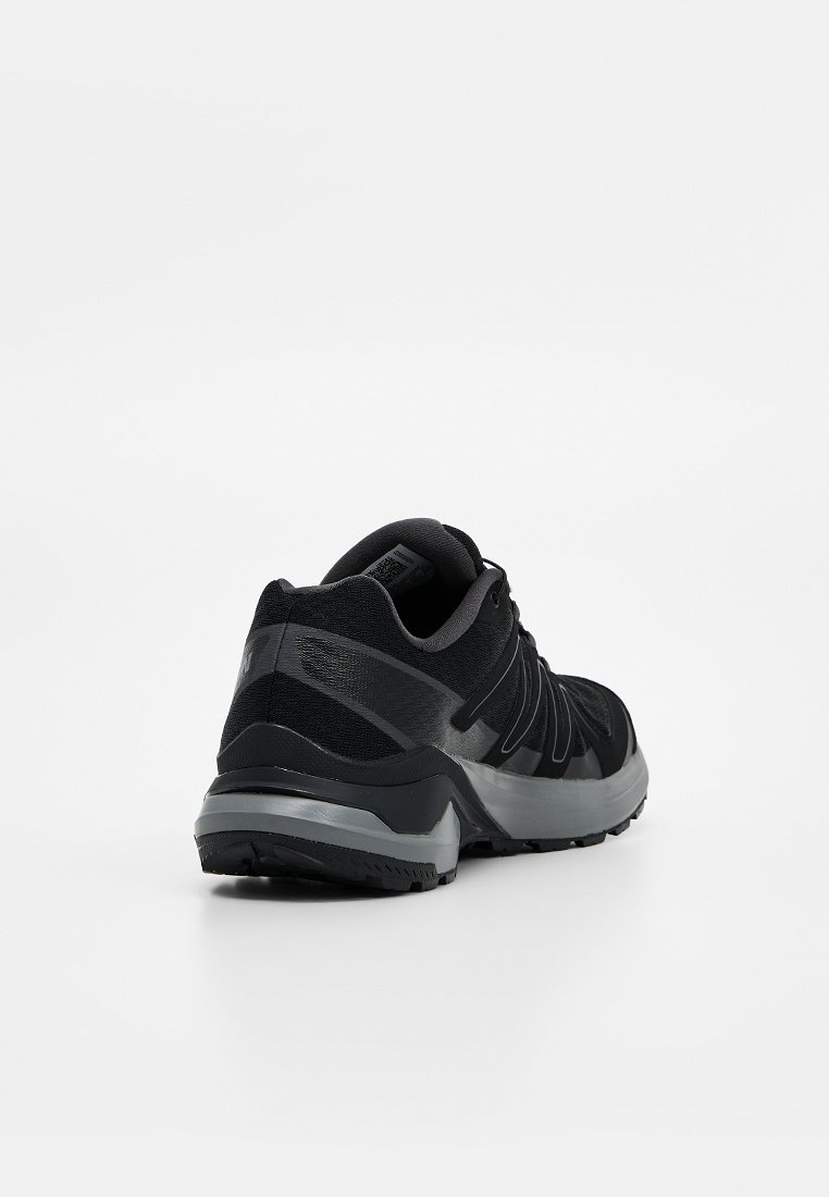 Zapatilla deportiva negra y gris, vista desde atrás y de lado, con suela texturizada y diseño con cordones sobre un fondo blanco liso.