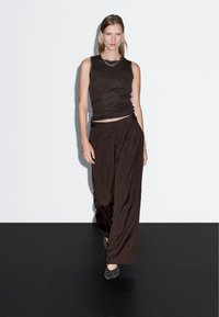 Brązowy top bez rękawów zestawiony z szerokimi, brązowymi spodniami. Gładki materiał z delikatnym drapowaniem. Modelka nosi także szare baleriny i srebrny naszyjnik z łańcuszka.