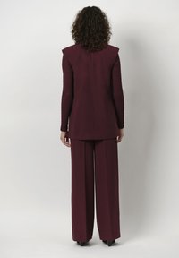 Bordeauxkleurig pak met een op maat gemaakt langer jasje met schoudervullingen en gestructureerde lange mouwen, gecombineerd met wijde broekspijpen.