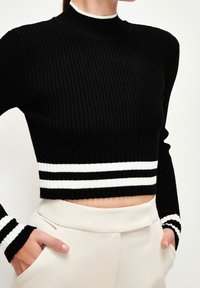 Pull cropped noir en tricot côtelé avec col montant, arborant des accents rayés blancs aux poignets et à l'ourlet. Associé à un pantalon de couleur claire.