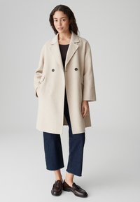 Vrouw in een beige dubbelzijdige jas, donkere cropped jeans, donkere loafers, staande tegen een effen grijze achtergrond.