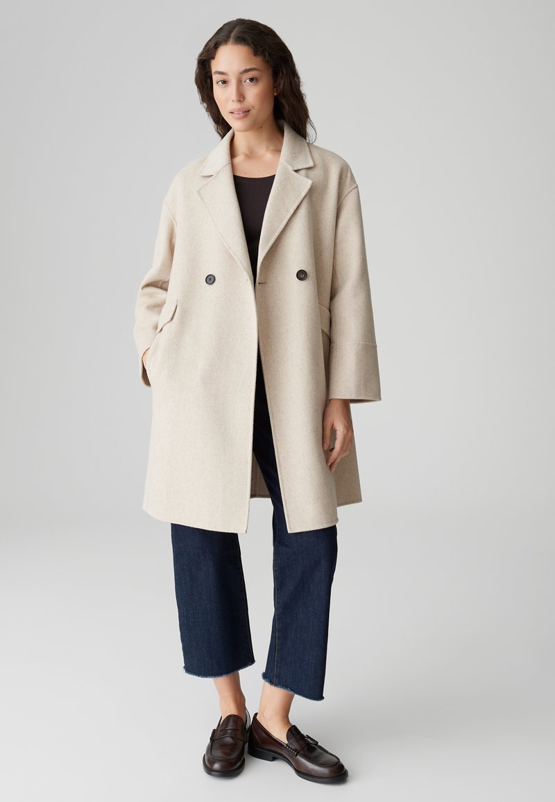 Vrouw in een beige dubbelzijdige jas, donkere cropped jeans, donkere loafers, staande tegen een effen grijze achtergrond.