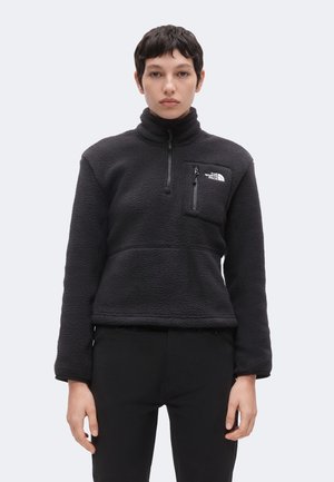 Osoba oblečená v čiernom fleecovom pulóvri so zipsovou náprsnou kapsou a logom The North Face, stojaca pred jednoduchým svetlým pozadím.