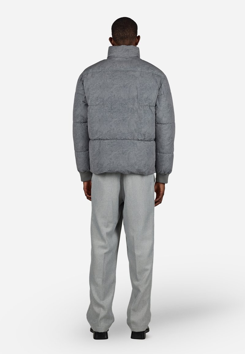 Hombre de pie de espaldas, vistiendo una chaqueta acolchada gris, pantalones gris claro y zapatos negros sobre un fondo blanco.