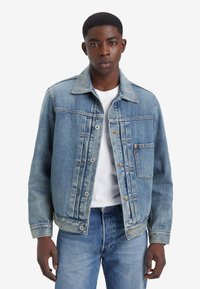 Jachetă din denim de culoare albastră deschisă, cu un sistem de închidere cu nasturi pe față, două buzunare la piept și un guler clasic, asortată cu un tricou alb și blugi tematici.