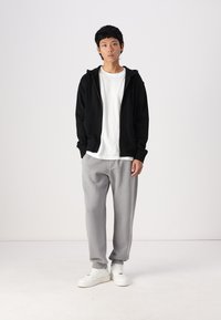 Sudadera negra con cremallera, camiseta blanca, pantalones de chándal grises y zapatillas blancas. El conjunto presenta un ajuste holgado y un diseño minimalista.