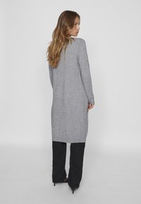 Cardigan long gris en tricot avec un ourlet et des poignets côtelés, présentant une coupe décontractée et une texture douce, associé à un pantalon noir à jambe large.