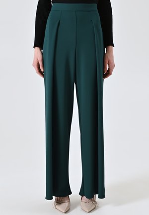 Pantaloni ampi in tessuto verde scuro, con pieghe frontali e una consistenza liscia. Abbinati a scarpe punta beige chiare.