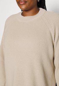 Beige neulepaita, jossa on ribbikuvio ja laaja pyöreä kaula-aukko. Raglan-hihat korostavat rentoa ja väljästi istuvaa leikkausta.