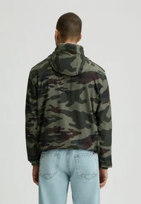 Zeleni, črni in burgundski kamuflažni jopič s kapuco; ima elastične manšete in ohlapen kroj, kombiniran s svetlo modrimi jeans hlačami.
