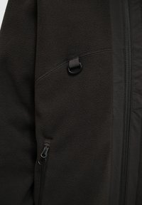 Mørk fleecejakke med en struktureret overflade, der har en lynlåslomme og en løkke detalje. Inkluderer kontrasterende stofpaneler.