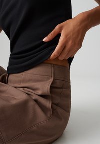 Zwart geribbeld shirt gelaagd over bruin op maat gemaakte broek met gedetailleerde stiksels; hand rustend op de taille, wat de textuur en pasvorm van de stof benadrukt.