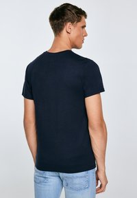 Noor mees küljelt vaates, seljas tavaline tume mereväe sinine lühikeste varrukatega t-särk ja helesinised teksad, ühtlase tausta ees.