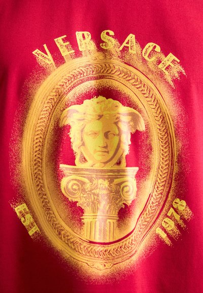 Raudonas audinys su auksine Medūzos galvos statula virš puošnios kolonos ovaliame rame, su tekstu „VERSACE“ viršuje ir „EST 1978“ apačioje.