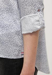 Biała koszula z granatowym, cętkowanym wzorem, podwiniętym mankietem z guzikiem i czerwonym akcentem. Na dole znajduje się czerwona i niebieska paskowana metka.