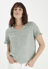 Grön V-ringad t-shirt med korta, fransade ärmar, tillverkad av ett mjukt, texturerat tyg med ett vitt abstrakt mönster över hela.