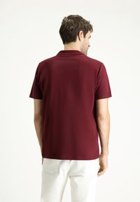 Polo shirt bordeaux con colletto classico, maniche corte e una texture liscia. Indossato con pantaloni bianchi, visto da dietro.