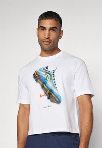 T-shirt bianca a maniche corte con un grafica colorata di un scarpino da calcio blu e giallo con dettagli neri e un logo Nike arancione sottostante.