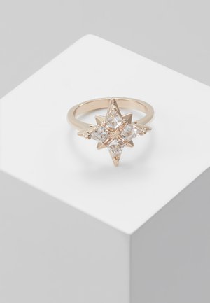 Kultasormus, jossa on neljä kirkasta, neliskanttista jalokiveä tähden muotoon aseteltuna, esillä valkoisella geometrisella jalustalla.