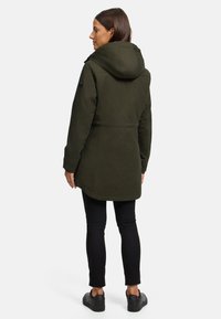 Manteau capuchon vert olive à la coupe décontractée, avec des accents de boutons et une texture lisse. Porté sur un jean skinny noir et des baskets foncées.