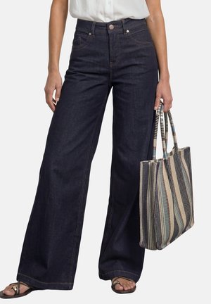 Person iført mørke, brede jeans og sandaler holder en stribet vævet taske, vist fra talje til fødder mod en ensfarvet baggrund.