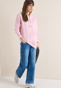 Femme portant un sweat à capuche rose, un jean bleu, des baskets blanches, tenant une veste foncée, debout devant un fond beige uni.